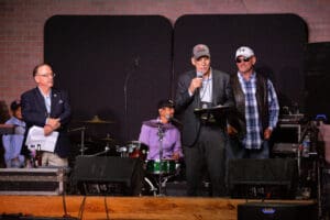 Men speaking on stage with band behind.