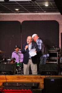 Man speaking on stage with microphone and papers.