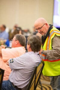 Man in safety vest talking to seated man.