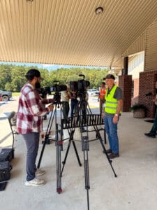 Cameraman filming man in safety vest outdoors.